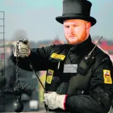Kominiarz w czarnym cylindrze i rękawicach trzyma szczotkę i obciążnik do czyszczenia komina.