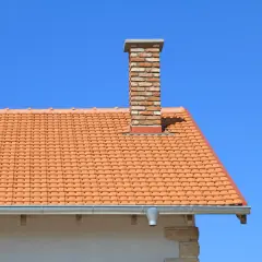 Ceglany komin do co na dachu pokrytym pomarańczową dachówką, na tle błękitnego nieba.