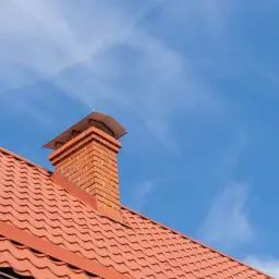 Ceglany komin na dachu z czerwonej dachówki, pod błękitnym niebem. Idealne rozwiązanie, czym obrobić komin na zewnątrz.