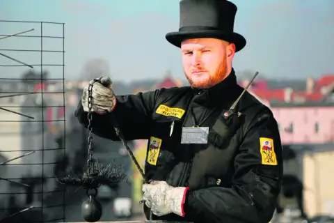 Kominiarz w czarnym cylindrze i rękawicach trzyma szczotkę i obciążnik do czyszczenia komina.