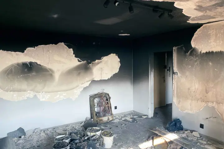 Spalony pokój z odpadającą farbą na ścianach i suficie. Widać zniszczony grzejnik i gruz. Pytanie: jak usunąć plamy na ścianie z komina?