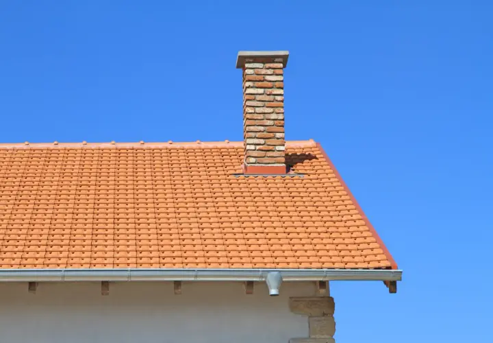 Ceglany komin do co na dachu pokrytym pomarańczową dachówką, na tle błękitnego nieba.
