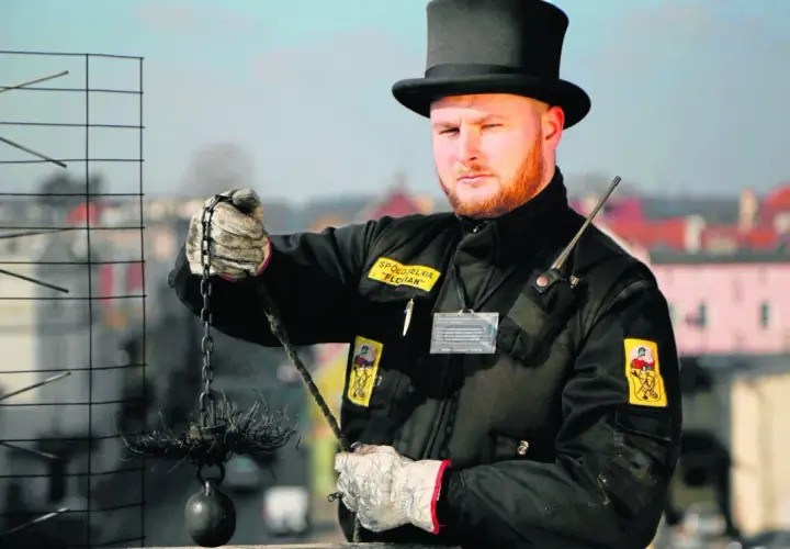 Kominiarz w czarnym cylindrze i rękawicach trzyma szczotkę i obciążnik do czyszczenia komina.
