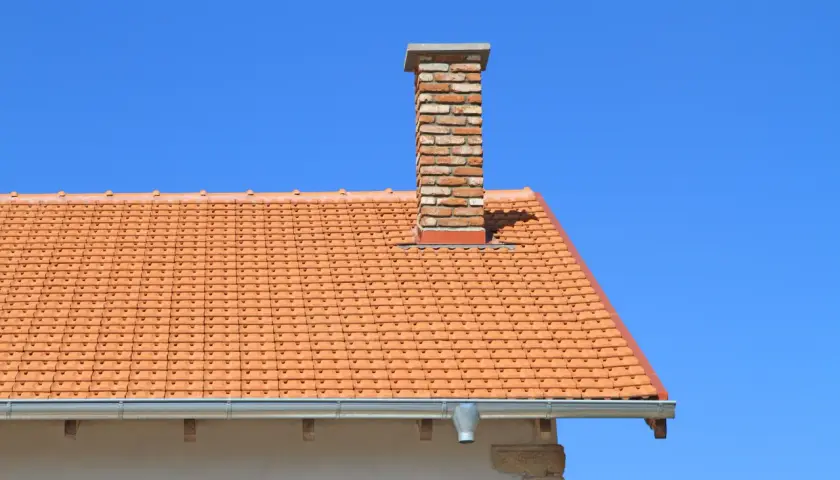 Ceglany komin do co na dachu pokrytym pomarańczową dachówką, na tle błękitnego nieba.