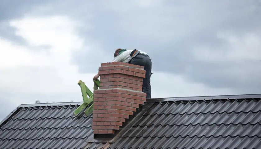 Budowa komina systemowego z cegły na dachu, pracownik w tle.