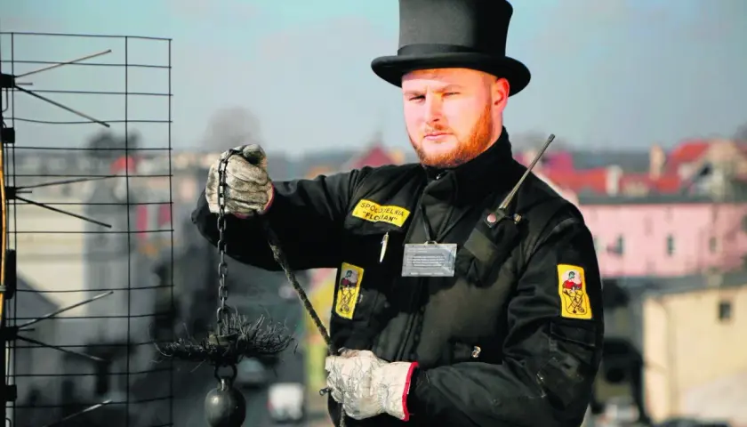 Kominiarz w czarnym cylindrze i rękawicach trzyma szczotkę i obciążnik do czyszczenia komina.