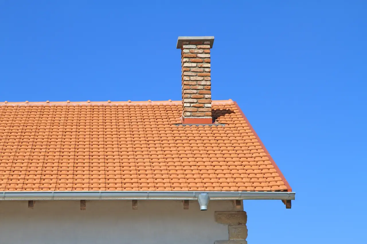 Ceglany komin do co na dachu pokrytym pomarańczową dachówką, na tle błękitnego nieba.